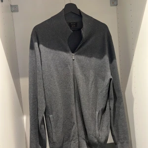 Grå tröja med dragkedja från Massimo Dutti - Snygg grå tröja från Massimo Dutti i exklusiv bomull. Tröjan har en dragkedja framtill och långa ärmar, perfekt för en stilren look. Passar bra till både vardag och mer uppklädda tillfällen. Storlek XL men Massimo Dutti har större storlekar så denna tröjan passar L