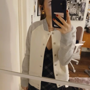 Bomberjacka från Under Armour - Snygg vit och grå bomberjacka från Under Armour. Jackan har en klassisk design med knappar framtill och långa ärmar. Perfekt för kyligare dagar med sin ColdGear-teknologi.