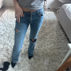 Blå lågmidjade jeans med slitningar - Säljer ett par blå jeans med slitningar på knäna. De har en klassisk femficksdesign och är perfekta för en avslappnad stil. Passar bra till en t-shirt eller hoodie.