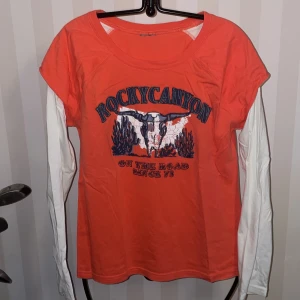 Orange t-shirt med tryck från Rocky Canyon - Cool orange t-shirt med tryck av en tjurskalle och texten 'On the road since 73'. Den har vita långa ärmar under de korta, vilket ger en lager-på-lager-look. Perfekt för en avslappnad stil.Storlek M-L