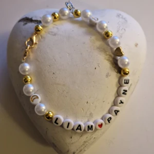 Pärlarmband med namn Liam Payne med hjärta - Charmigt armband med vita och guldiga pärlor samt bokstavspärlor som bildar namnet 'LIAM PAYNE'. Armbandet har hjärtdetaljer och en liten hjärtberlock. Perfekt för fans som vill minnas. 
