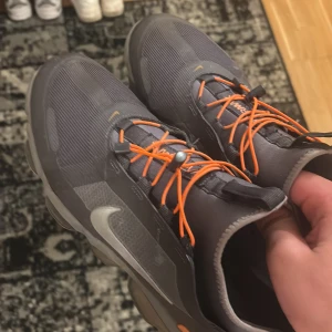 Nike sneakers med orange detaljer - Snygga grå Nike sneakers med orange snörning och detaljer. Skorna har en unik sula med synliga luftkuddar för extra komfort. Perfekta för både träning och vardag. Nypris 3000