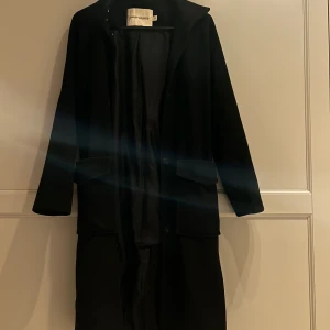 Svart kappa från Carin Wester - Elegant svart kappa från Carin Wester med knappar och knytskärp i midjan. Perfekt för kyligare dagar med sin långa design och klassiska stil.