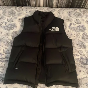 Svart dunväst från The North Face - Säljer en svart dunväst från The North Face med dragkedja och broderad logga på bröstet. Västen har en hög krage och är perfekt för kyligare väder. Modellen är klassisk och stilren. PASSAR SOM XS 