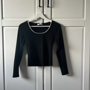 Svart långärmad topp med pärldetaljer från Mango💕 - Elegant svart långärmad topp från Mango med pärldetaljer runt halsringningen. Perfekt för en stilren look. Tillverkad i ett ribbat material som ger en fin struktur. Älskarrr!!❤️