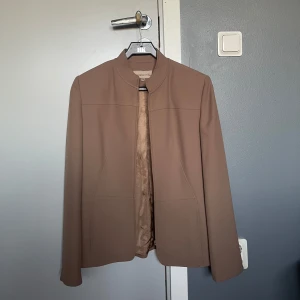 Kavaj - En elegant kavaj med en stilren design. Färgen är lite mörkare än vad den är på bilden. (lite mörk beige/oliv/brun färgad). Svårt att förklara färgen.  Kavajen har långa ärmar och en enkel, klassisk passform. Perfekt för en stilren look. Storlek 38. 