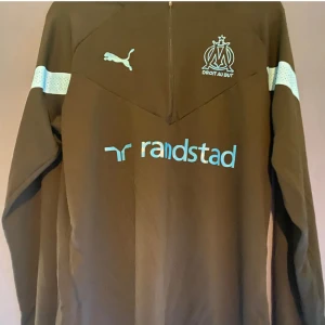 Marseille långärmad tröja från Puma - Säljer en Marseille långärmad tröja från Puma med ljusblå detaljer och logotyp. Tröjan har vita band på ärmarna och en tryckt text på framsidan. Perfekt för sportiga tillfällen!