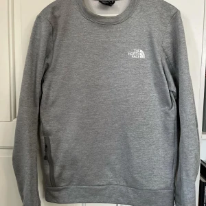 Grå sweatshirt från The North Face - Snygg och bekväm grå sweatshirt från The North Face med rund hals och långa ärmar. Perfekt för kyligare dagar och har en diskret logga på bröstet. Passar till både vardag och avslappnade tillfällen.