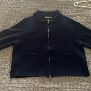 Mörkblå cardigan med dragkedja - Snygg mörkblå cardigan med dragkedja och två stora fickor framtill. Perfekt för en stilren look. Jackan har en klassisk krage och är tillverkad i ett mjukt material. Den har ett litet hål vid kragen (se sista bilden) men inget man tänker på vid användning, det går även att sy ihop lätt med nål och tråd! Köpt för 600kr på Åhléns men pga av hålet säljer jag för 300kr 