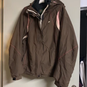 Brun jacka från Peak Performance - Säljer en brun jacka från Peak Performance med rosa och vita detaljer. Jackan har en dragkedja och är långärmad, perfekt för höst och vinter. Den har en logotyp på bröstet och en praktisk huva.