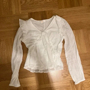 Vit blus från Gina Tricot - Säljer en söt vit blus från Gina Tricot med volangdetaljer och v-ringning. Blusen har långa ärmar och en lätt rynkad design som ger en romantisk känsla. Perfekt för både vardag och speciella tillfällen.