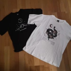 Svart och vit t-shirt från 157 - Två t-shirts från 157 i storlek S. Den svarta t-shirten har ett tryck med texten 'Stay in the moment' och en grafisk design med sol och måne. Den vita t-shirten har ett tryck med en orm och texten 'Different perspective'. Ni kan köpa en T-shirt för 25kr och båda för 40kr