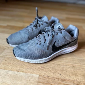 Grå Nike löparskor - Säljer ett par grå Nike löparskor med svart logga. Skorna har snörning och en bekväm sula för optimal löpning. Perfekta för träning och vardagsbruk.