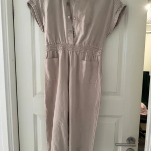 Beige jumpsuit med knappar - Snygg beige jumpsuit med korta ärmar och knappar framtill. Den har en elastisk midja och praktiska fickor. Perfekt för en avslappnad stil. Lappen är kvar. 