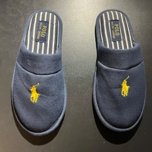 Polo Ralph Lauren tofflor blåa - Sköna blå tofflor från Polo Ralph Lauren med en gul broderad logotyp på ovansidan. Innersulan har ett randigt mönster i blått och vitt. Skick 7,5/10 har 4 små defekter på innersidan, från min hund 🐶 (se på bild 4).   Perfekta för att ha hemma. Nypris ca 500-600 kr. Priset är inte hugget i sten 🪨. Skriv om ni är intresserade.  Mvh Sigge🤙🥂