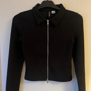 Svart ribbad jacka från H&M Divided - Snygg svart ribbad jacka från H&M Divided med dragkedja framtill. Jackan har en klassisk krage och är croppad i modellen. Perfekt för en stilren look.