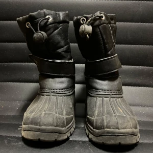 Svarta vinterstövlar från 157 - Säljer ett par svarta vinterstövlar från 157 med robust sula och justerbar rem. Perfekta för kalla dagar med bra grepp och värmeisolering. Stövlarna har en praktisk dragsko upptill för extra skydd mot snö och kyla.