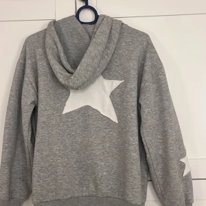 Grå hoodie med stjärnor från Gina Tricot - Säljer en grå hoodie från Gina Tricot med dragkedja och stor vita stjärnor på ryggen och en liten på armen. Väldigt gosig🩶