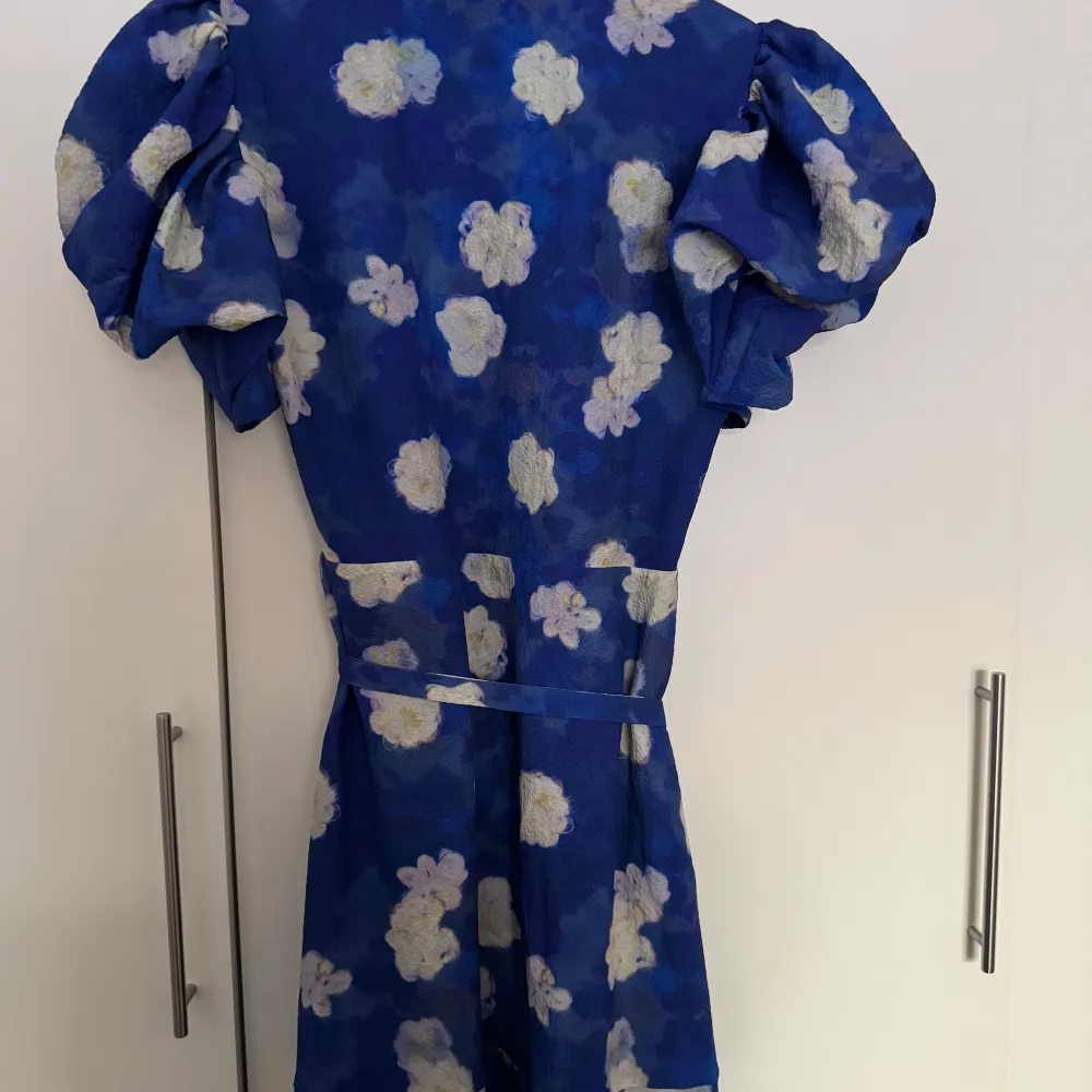 Snygg blå omlottklänning med blommigt mönster. Klänningen har korta puffärmar och ett knytband i midjan för en smickrande passform. Använd en gång i Italien. Storlek 38.. Mekot.