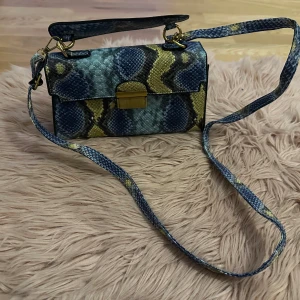 Ormmönstrad axelväska med gulddetaljer - Snygg axelväska med ormmönster i blått och gult. Väskan har en justerbar axelrem och ett praktiskt spänne i guld. Perfekt för att ge din outfit en trendig touch.