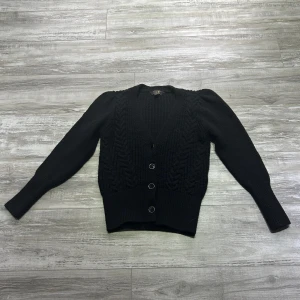 Svart stickad kofta från Lipsy - Snygg svart stickad kofta från Lipsy med flätstickat mönster och knappar framtill. Koftan har långa ärmar och en v-ringad design, perfekt för kyligare dagar. Använd 2 gånger.