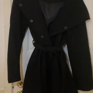 Svart kappa med bälte - Elegant svart kappa med knappar och bälte i midjan. Perfekt för kyligare dagar med sin långa design och stilrena look.