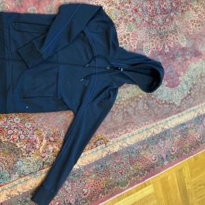 Mörkblå huvtröja från BOSS - Säljer en stilren mörkblå huvtröja från BOSS med dragkedja och justerbar huva. Tröjan har långa ärmar och subtila blå detaljer längs sömmarna. Perfekt för en avslappnad look. 