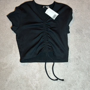 Svart ribbad topp från H&M - Snygg svart ribbad topp från H&M med korta ärmar och v-ringning. Toppen har en trendig snörning framtill som ger en fin rynkad effekt. Perfekt för en stilren look. Toppen är oanvänd!