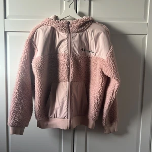 Rosa teddyjacka från Champion - Mysig rosa teddyjacka från Champion med huva och dragkedja. Perfekt för kyliga dagar med sin fluffiga textur och bekväma passform. Jackan har långa ärmar och ribbade muddar för extra komfort. Så söt och bekväm! 