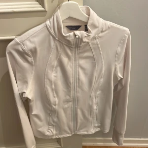 Vit yoga jacka från Danskin - Säljer en vit yoga/pilates/tränings jacka från Danskin med dragkedja framtill. Jackan har en hög krage och långa ärmar, perfekt för träning eller vardagsbruk. Den är tillverkad i ett syntetmaterial som ger en bekväm passform.