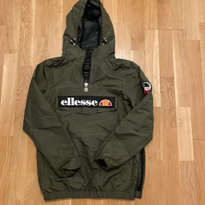 Grön vindjacka från Ellesse - Snygg grön vindjacka från Ellesse med huva och dragkedja. Jackan har ett stort Ellesse-märke på framsidan och en praktisk ficka. Perfekt för blåsiga dagar.