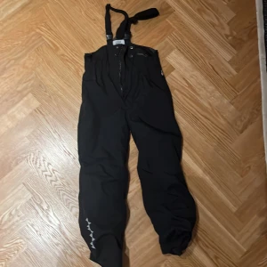 Svarta skidbyxor från Isbjörn of Sweden - Säljer ett par svarta hängselbyxor från Isbjörn of Sweden. Byxorna är vattentäta och har en andningsförmåga på 15,000g/m2/24h. Perfekta för utomhusaktiviteter i alla väder. De har justerbara hängslen och är designade för att hålla kroppen torr och varm.
