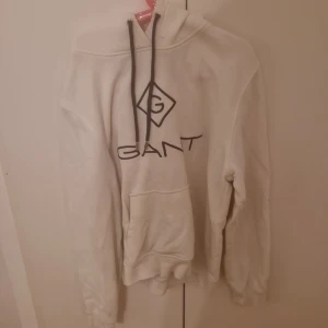 Vit hoodie från Gant - Säljer en stilren vit hoodie från Gant med svart logotyp på bröstet. Hoodien har en klassisk känguruficka och justerbar dragsko i huvan. Perfekt för en avslappnad look.