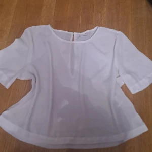 Vit blus från Gina Tricot - Säljer en stilren vit blus från Gina Tricot i storlek M mrn passar dom S. Blusen har korta ärmar och en rund halsringning med en liten knappdetalj i nacken. Perfekt för en enkel och elegant look.