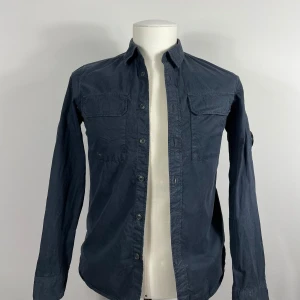 Mörkblå bomullsskjorta - Snygg mörkblå skjorta i 100% bomull med långa ärmar och knappar framtill. Perfekt för en stilren look. Köpt på Zgander i Helsingborg. Modelldockan är lite större än va plagget är vilket gör att den ser lite konstigt formad ut. 