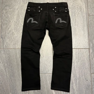 Svarta jeans från Evisu - Snygga svarta jeans från Evisu med unika detaljer på bakfickorna. Längd 95cm bredd 44cm fotvidd 19cm