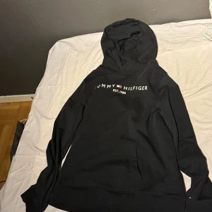 Svart hoodie från Tommy Hilfiger - Säljer en stilren svart hoodie från Tommy Hilfiger med logotyp och text 'EST. 1985' på bröstet. Tröjan har en klassisk huva och långa ärmar, perfekt för en avslappnad look.