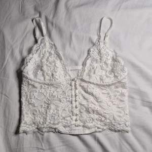 Vit spetslinne med pärlknappar - Säljer en vacker vit spetslinne med justerbara axelband och dekorativa pärlknappar framtill. Toppen har en romantisk känsla med sitt detaljerade spetsmönster och är perfekt för speciella tillfällen.