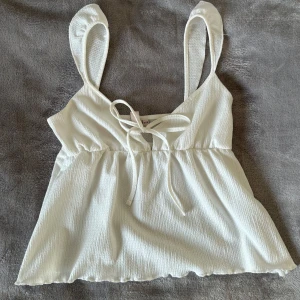 Vit topp från Nelly - Säljer en söt vit linnetopp från Nelly med knytband framtill. Toppen har tunna axelband och en lätt rynkad effekt i tyget. Perfekt för varma sommardagar! Använd 1 gång, storlek xxs. Bra skick