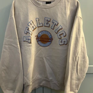 Beige sweatshirt från Gina Tricot - Säljer en beige sweatshirt från Gina Tricot med trycket 'Athletics Madison Wisconsin United States' i blått och rött. Tröjan har långa ärmar och ribbade muddar vid hals, ärmslut och nederkant.