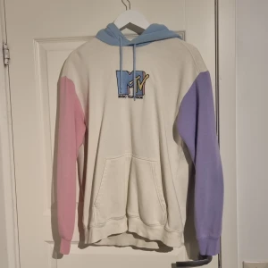 MTV hoodie med pastellfärger - Snygg hoodie från MTV med en cool logga på framsidan. Tröjan har en beige bas med kontrasterande ärmar i rosa och lila samt en ljusblå huva. Perfekt för en avslappnad stil.