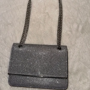 Glittrig silver axelväska - Snygg och glittrig axelväska i silver med kedjerem. Perfekt för att ge en extra touch till din outfit. Väskan har en stilren design och är lätt att bära med sig.