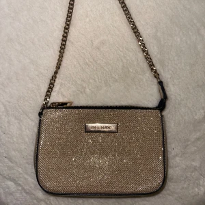 Glittrig axelväska från River Island  - Snygg glittrig axelväska från River Island med en kedjerem och dragkedja. Perfekt för att ge en touch av glamour till din outfit. Väskan är i en elegant guldton och har en praktisk storlek för det viktigaste.