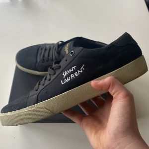 Svarta sneakers från Saint Laurent - Snygga svarta sneakers från Saint Laurent med vit logga på sidan. Skorna har en klassisk design med snörning och en beige sula. Perfekta för en stilren look. Hör av er om ni har fler frågor