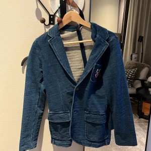 Blazer från Ralph lauren - Snygg blå kavaj och två framfickor. Kavajen har knappar framtill och en broderad detalj på bröstet. Perfekt för en stilren look med en twist.