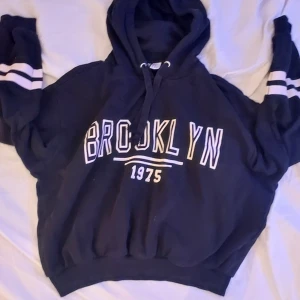 Svart hoodie från H&M Divided - Säljer en svart hoodie från H&M Divided med texten 'Brooklyn 1975' på framsidan. Tröjan har en huva med dragsko och vita ränder på ärmarna. Perfekt för en avslappnad stil.