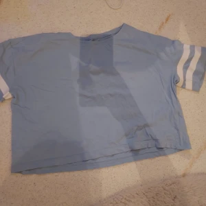 Blå kortärmad topp från H&M - Säljer en ljusblå kortärmad topp från H&M i ekologisk bomull. Toppen har vita ränder på ärmarna och en avslappnad passform. Perfekt för en casual look.