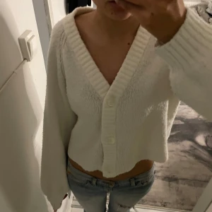 Vit stickad kofta från Gina Tricot - Mysig vit stickad kofta från Gina Tricot med v-ringning och knappar framtill. Perfekt för kyliga dagar och ger en stilren look. Koftan har långa ärmar och en croppad passform som passar bra till jeans eller kjol.