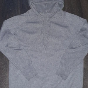 Grå merino ull hoodie  - Säljer en stilren grå stickad hoodie med långa ärmar och ribbade muddar. Perfekt för kyliga dagar med en bekväm passform och justerbar huva. Ett måste i garderoben för en avslappnad look.