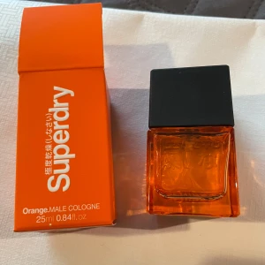 Superdry Orange Male Cologne - Säljer en fräsch och energisk herrparfym från Superdry. Flaskan är i en djärv orange färg med en stilren svart kork. Förpackningen matchar med samma livfulla orange ton. Perfekt för den som vill ha en uppfriskande doft.